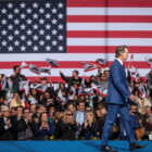 California Governor Gavin Newsom walks across a stage during his inauguration in January 2023. The lower half of the back drop is fuzzy images of people waving flags and the upper half is a giant US flag.