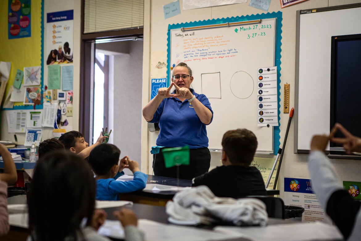 Teacher in a blue polo uses hands to demonstrate shapes to a small group of 5-year-olds